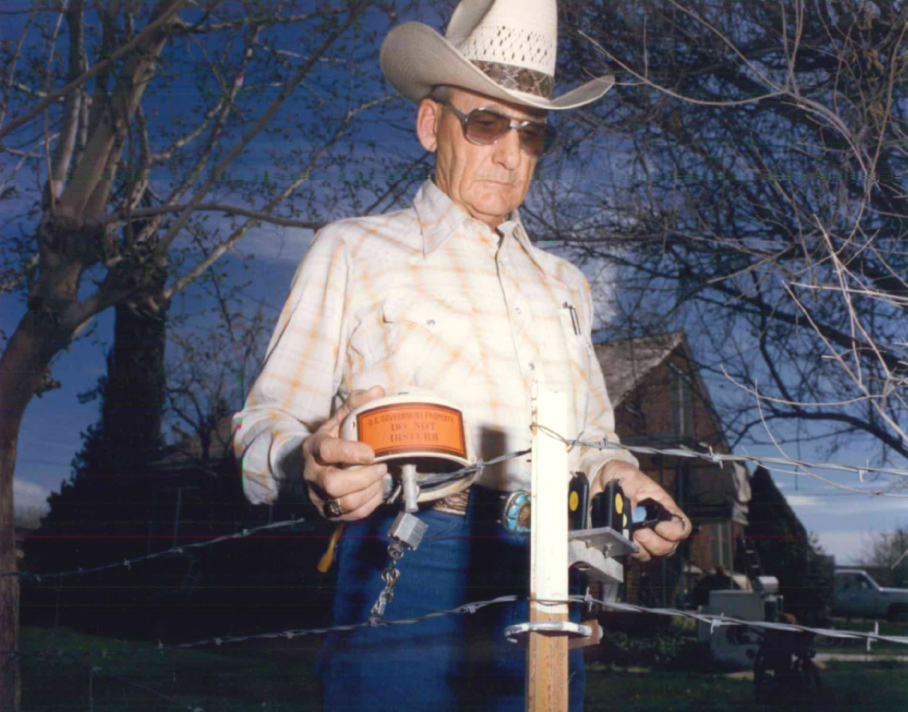 An older man wearing a cowboy hat, sunglasses, and a mustache, holds a device near a barbed wire fence.