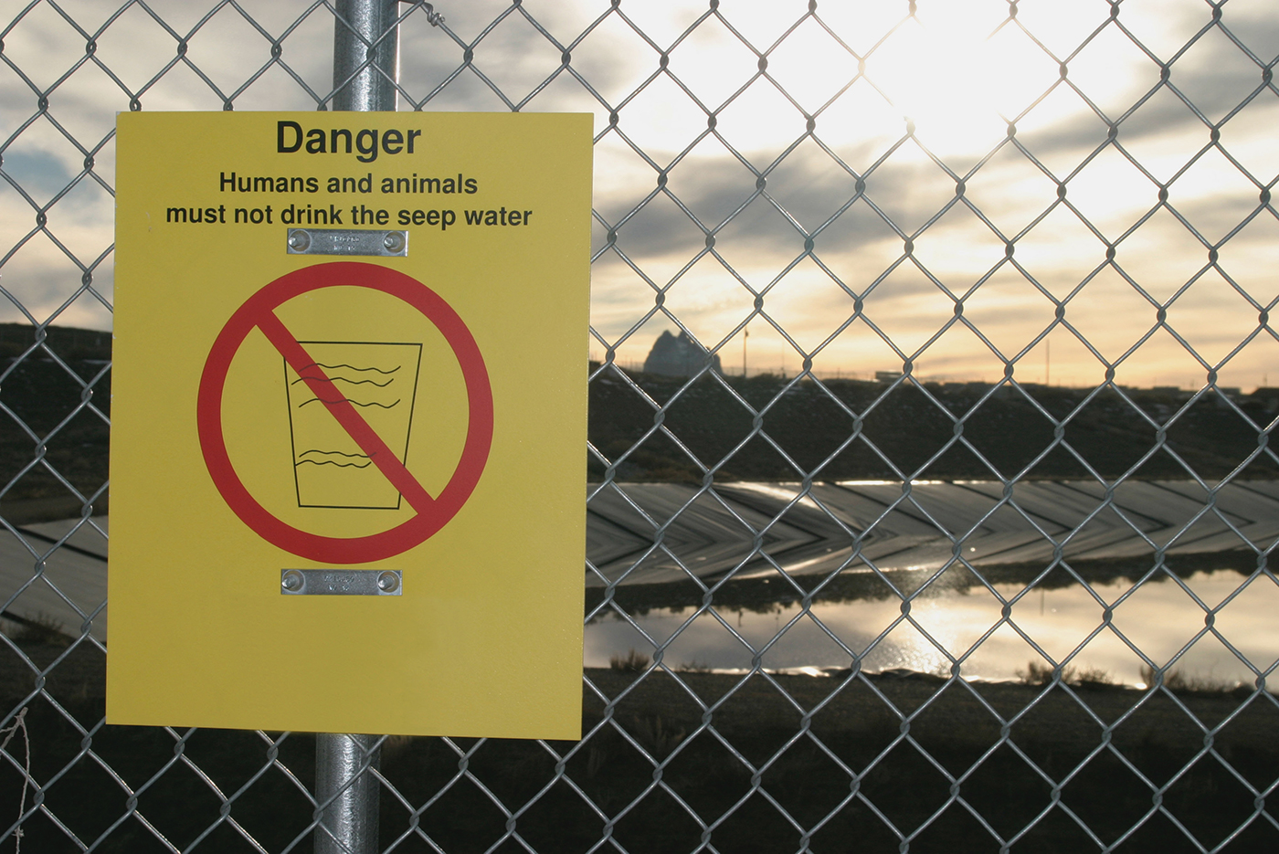 A yellow sign hangs on a wire fence in front of a reservoir. The sign reads, 'Danger: Humans and animals must not drink the seep water'. The sign depicts a crossed out glass of water.
