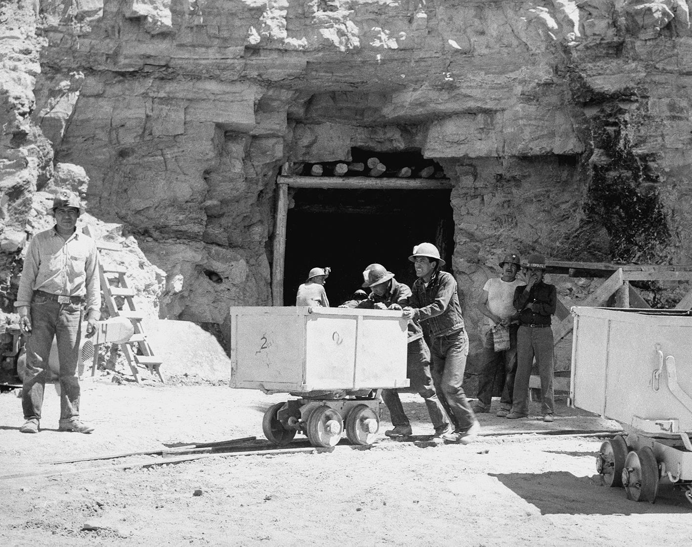 Men push a cart along a track. The entrance to a mine can be seen in the background.