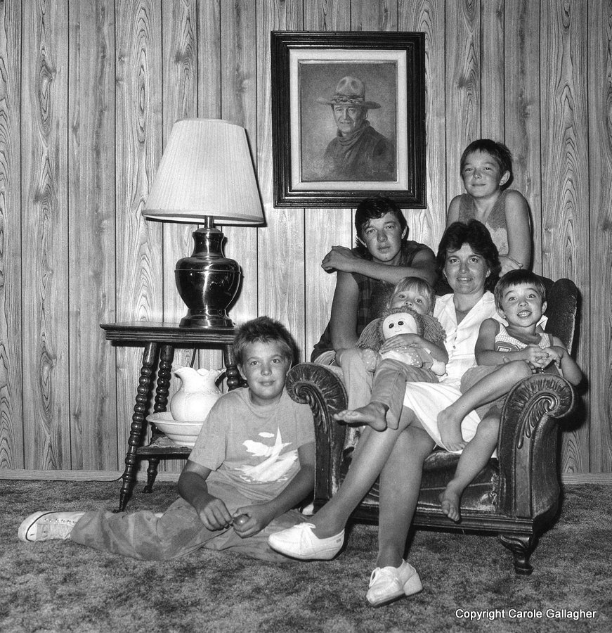 A woman sits in an armchair surrounded by her five children. A portrait of a man in a cowboy hat hangs on the wood-paneled wall.