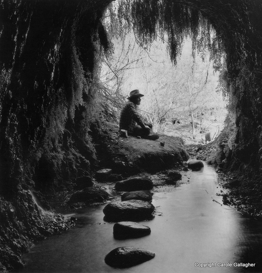 A man in a stetson sits on a rock near the mouth of a small cave with a stream running through it.
