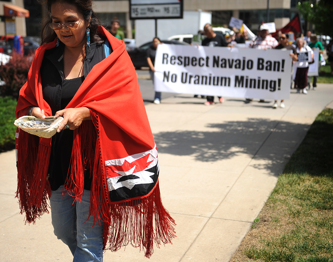 A woman wearing a patterned red blanket carries a shell full of sage branches. A small crowd carries a sign that reads, "Respect Navajo Ban! No Uranium Mining!".
