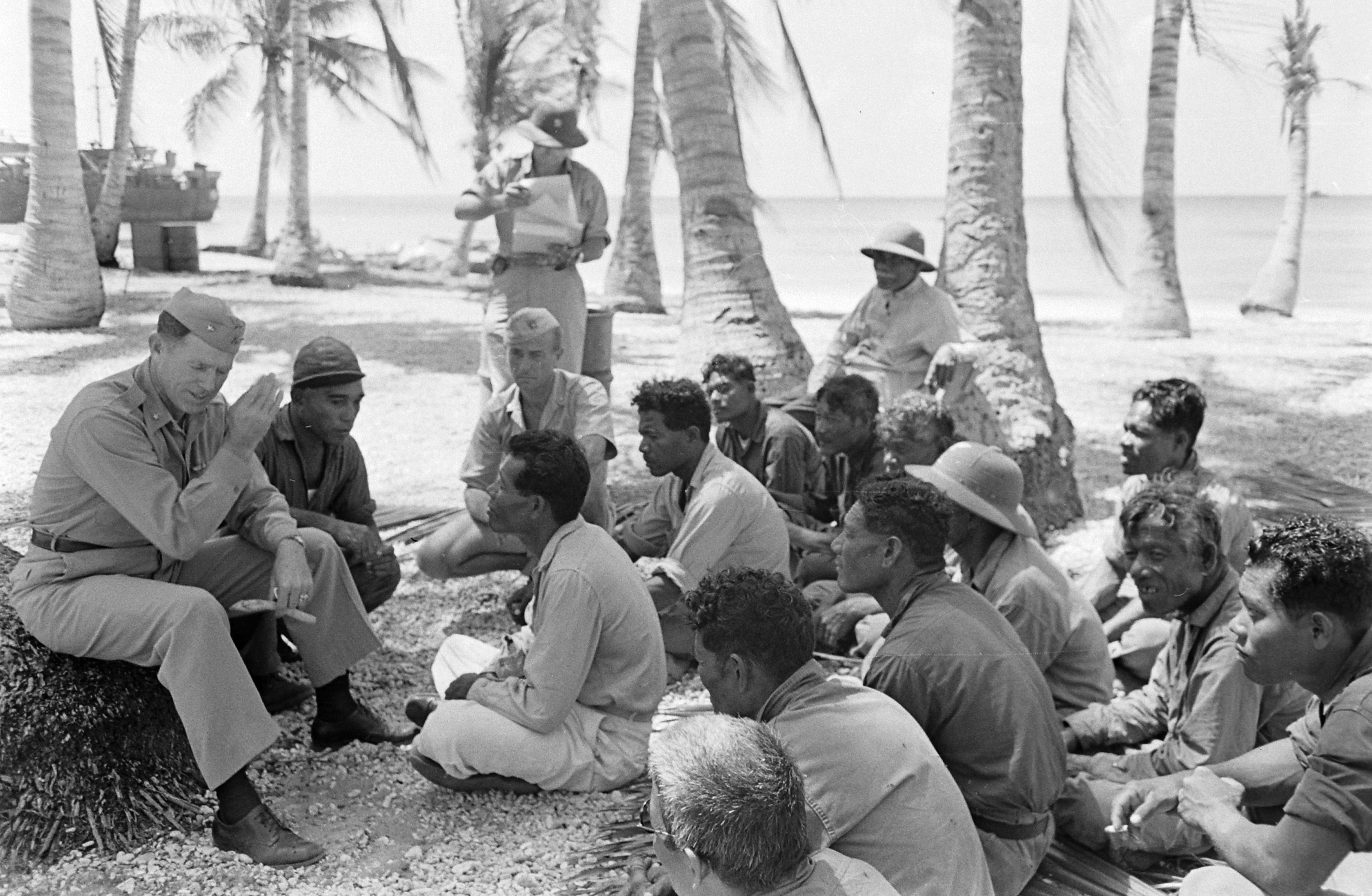 A military-uniformed man sits on a log and talks to a group of men sitting cross-legged on a rocky beach.