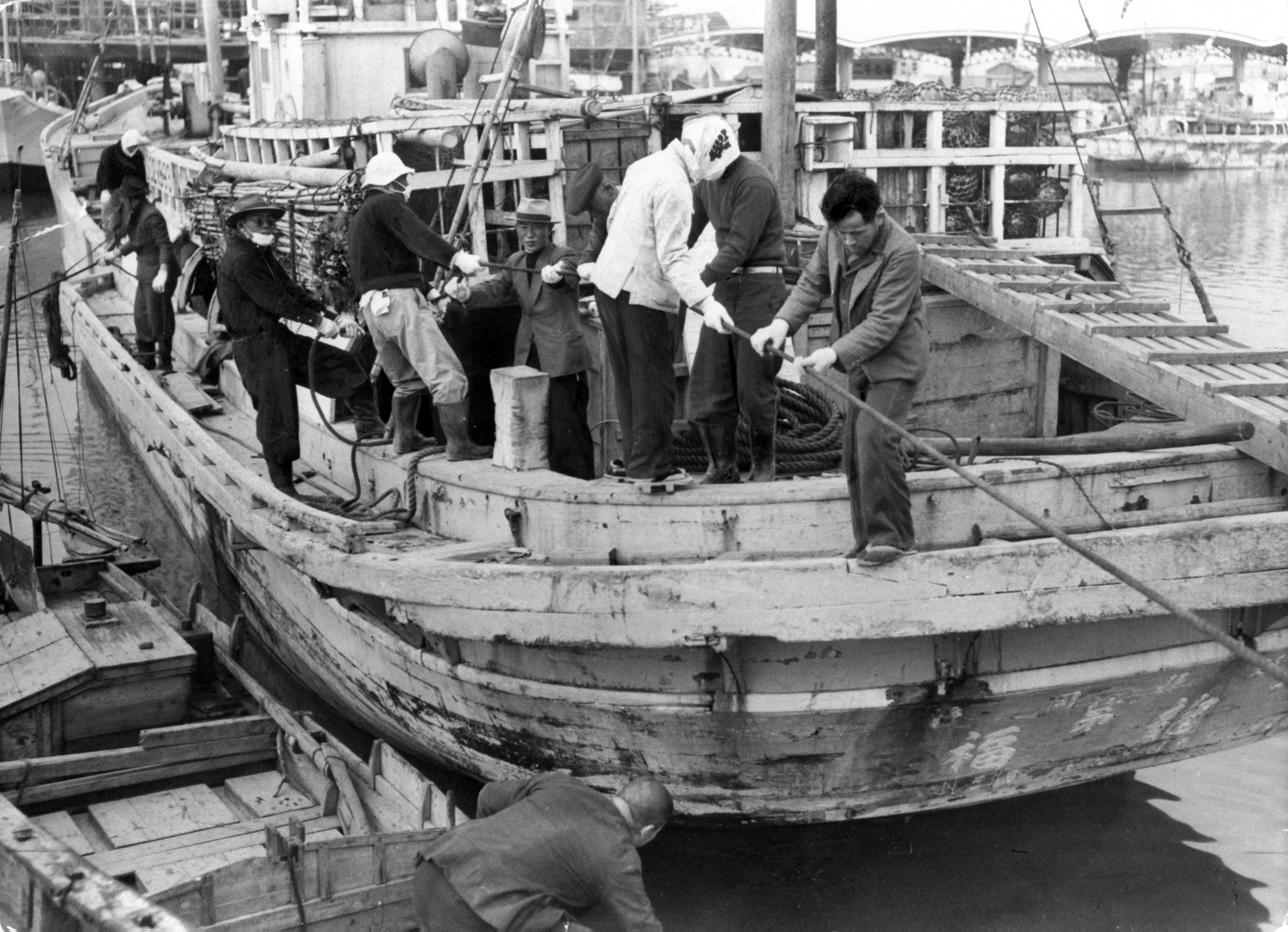 People with face-masks and gloves inspect a wooden fishing boat as it's docked near a pier.