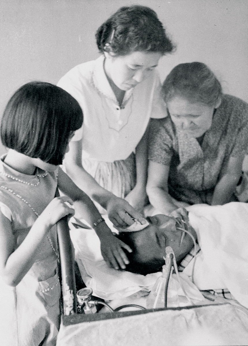 A girl, a woman, and an older woman stand over a man laying sick in bed. The man has tubes for breathing coming out of his nose. The woman wipes his forehead of sweat.