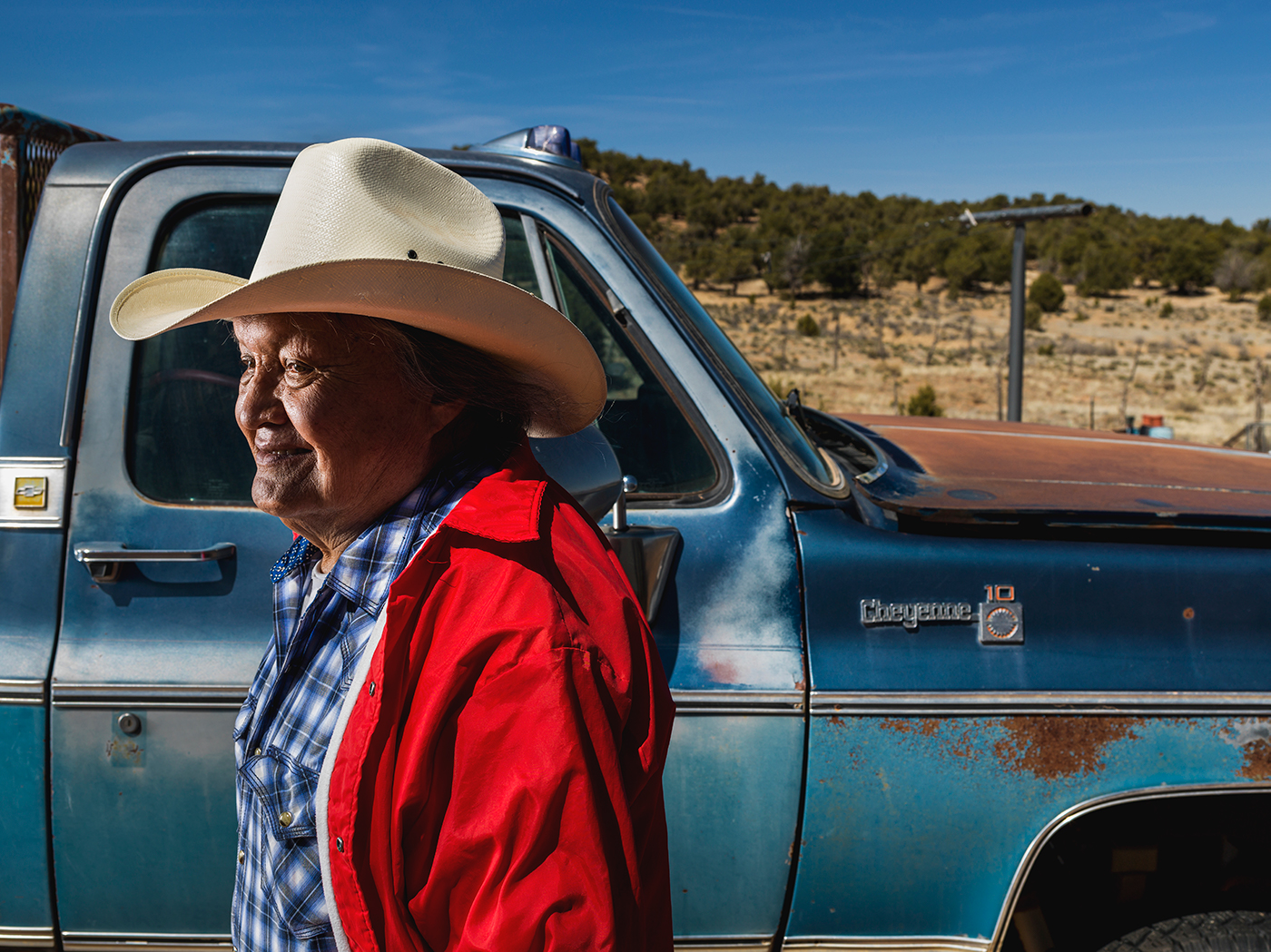 A man wearing a blue patterned button down, a red sip-up hoodie, and a cowboy hat, is seen in front of a rusted blue truck.