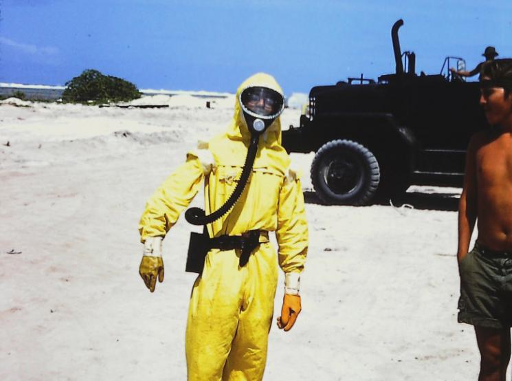 A young man wearing a yellow radiation suit and face mask stands on a beach near a shirtless man wearing a ball-cap.