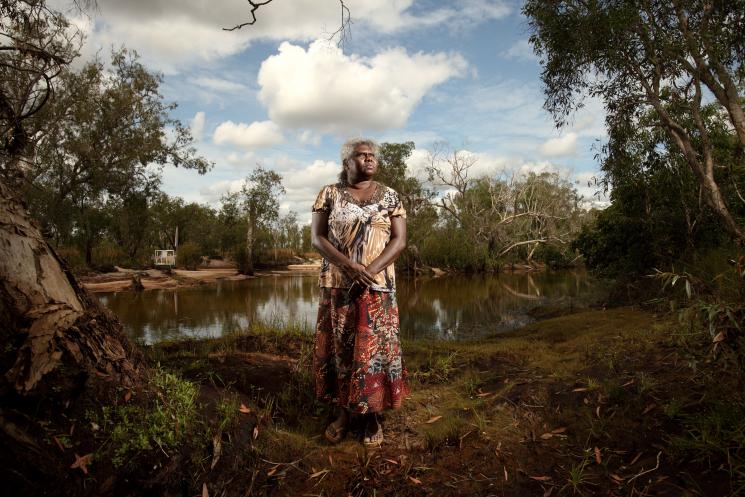A woman in a patterned shirt and skirt stands in a clearing near a river.