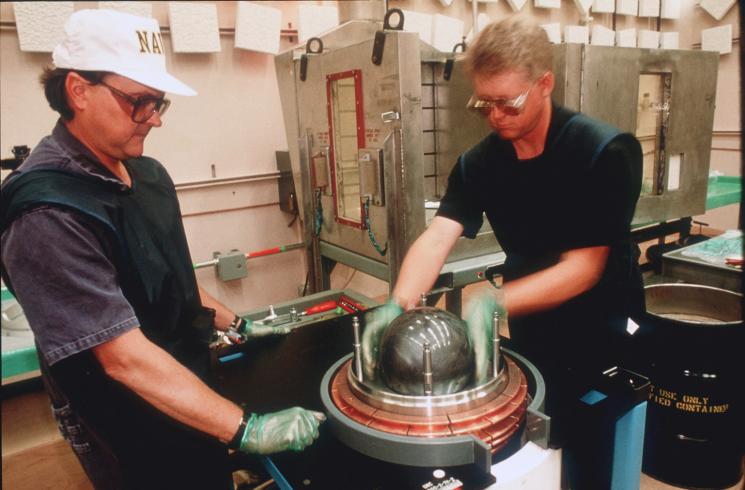 Two workers wearing plainclothes and green safety vests lift a grey sphere out of a device in a lab-like room.