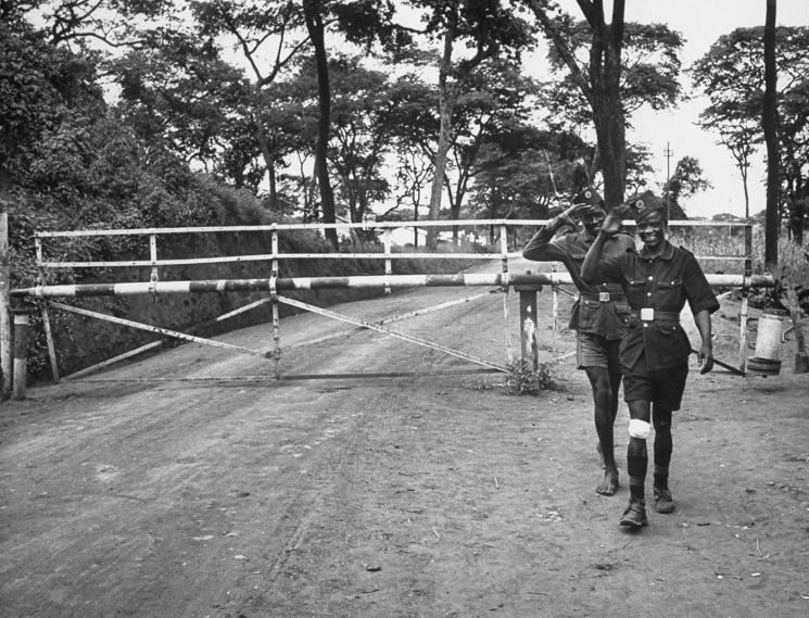 Two men in short-sleeve uniforms stand on a dirt road in front of a closed gate. 