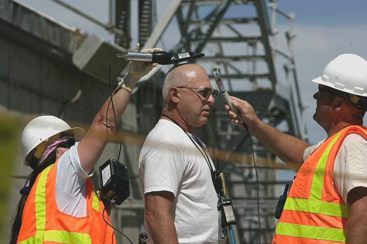 Two workers in reflective orange and yellow vests and hard hats wave instruments over a bald man wearing sunglasses.