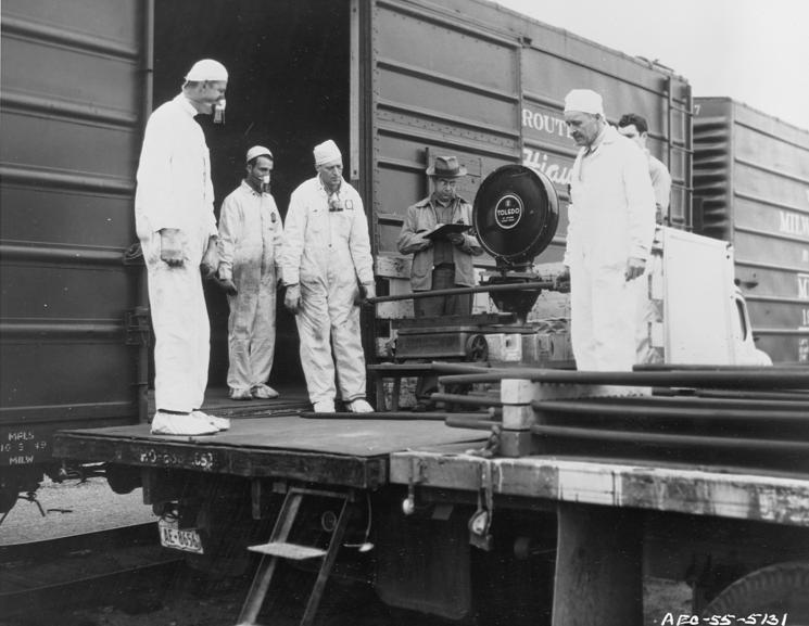 People in white jumpsuits and respirators stand around a scale next to a railcar.