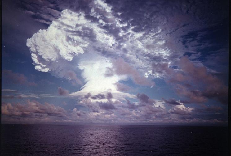 An explosion and mushroom cloud light up a dark purple sky, over a calm ocean.