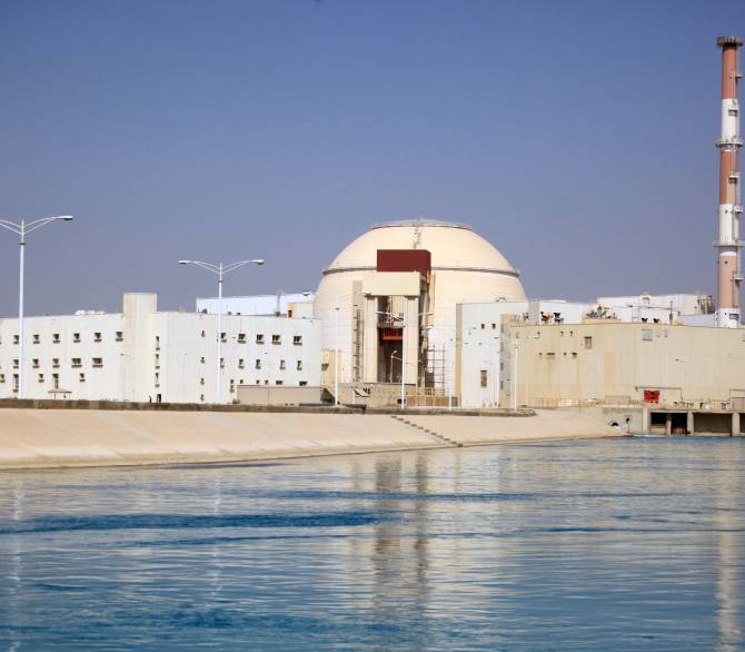 A tan and white domed building with a smokestack sits behind a body of water.