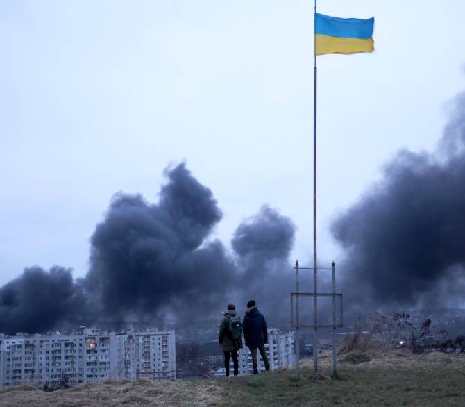 People standing near a Ukrainian national flag watch as dark smoke billows following an air strike in the western Ukrainian city of Lviv, on March 26, 2022.