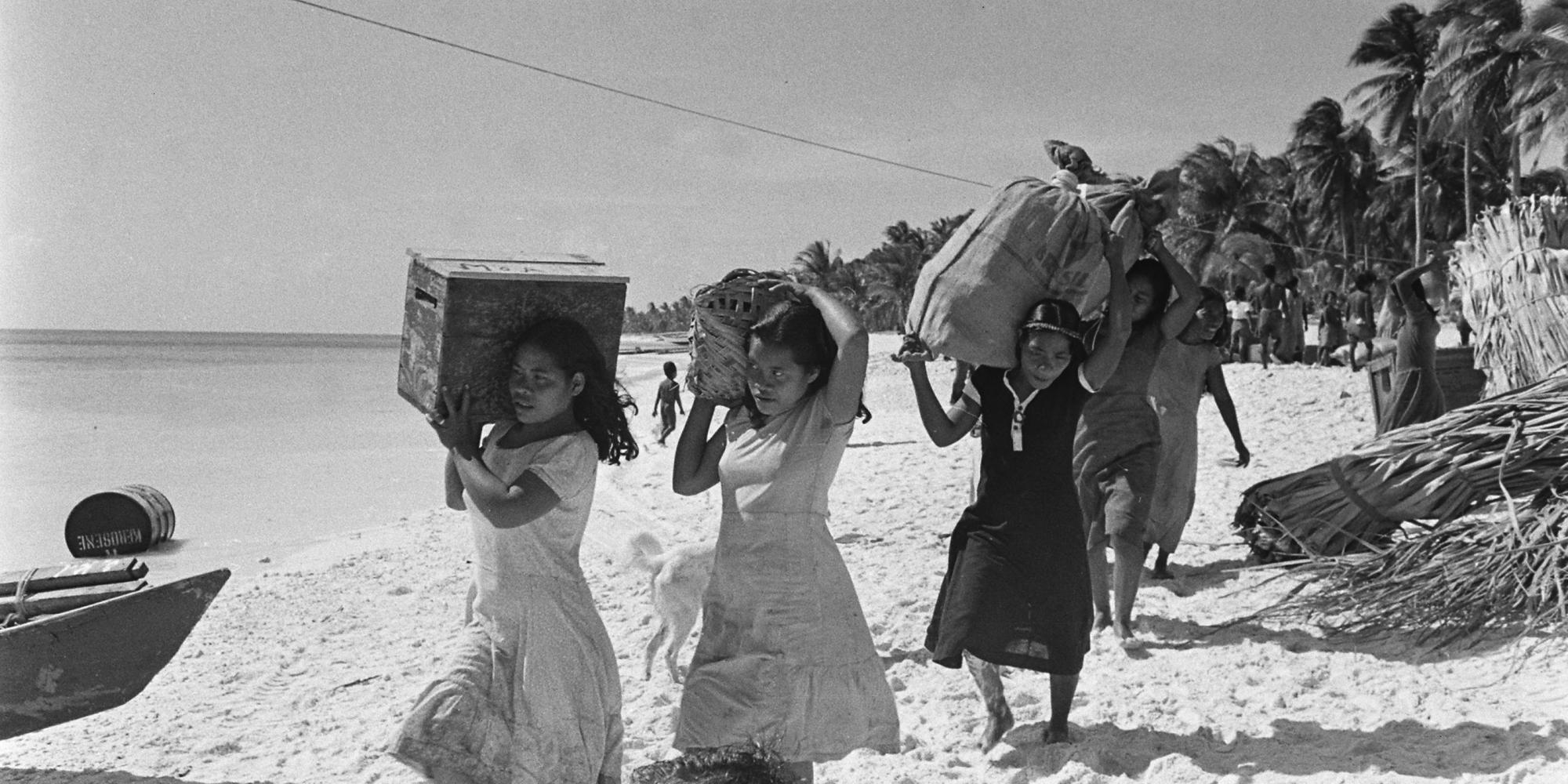 A group of women carry boxes and bags across a beach.